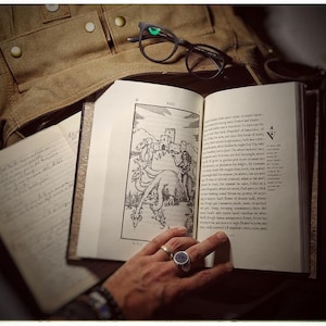K&ouml;nnte beinhalten: Ein offenes Buch mit einer Schwarzwei&szlig;illustration eines Drachen und einer Burg. Das Buch liegt auf einer braunen Lederoberfl&auml;che. Eine Hand mit einem Ring h&auml;lt das Buch offen.