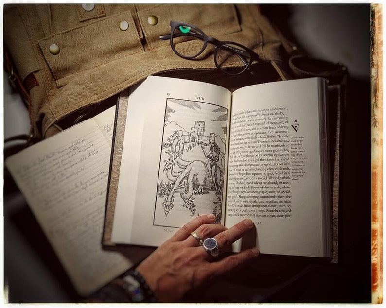 K&ouml;nnte beinhalten: Ein offenes Buch mit einer Schwarzwei&szlig;illustration eines Drachen und einer Burg. Das Buch liegt auf einer braunen Lederoberfl&auml;che. Eine Hand mit einem Ring h&auml;lt das Buch offen.