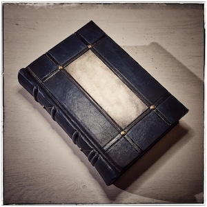 K&ouml;nnte beinhalten: Ein dunkelblaues, ledergebundenes Notizbuch mit einem rechteckigen, cremefarbenen Mittelteil. Der Einband ist mit dunklen Linien und kleinen, goldfarbenen Nieten verziert. Der Buchr&uuml;cken hat ein abgerundetes, strukturiertes Design.