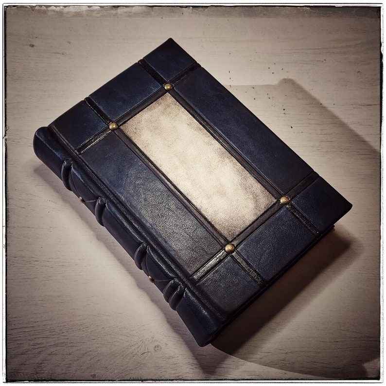 K&ouml;nnte beinhalten: Ein dunkelblaues, ledergebundenes Notizbuch mit einem rechteckigen, cremefarbenen Mittelteil. Der Einband ist mit dunklen Linien und kleinen, goldfarbenen Nieten verziert. Der Buchr&uuml;cken hat ein abgerundetes, strukturiertes Design.