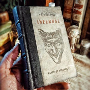 Könnte beinhalten: Ein Buch im Vintage-Stil mit dem Titel "Infernal" und einer Teufelsgesicht-Illustration auf dem Cover. Das Buch hat einen schwarzen Buchrücken und wird in einer Hand gehalten. Der Titel ist auf Französisch.