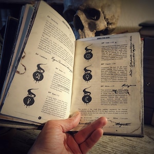 Op de afbeelding: Een open boek met een schedel erop. Het boek is getiteld "Advanced Potion Making" en heeft een sectie over ketels. De tekst beschrijft de verschillende soorten ketels en hun eigenschappen, waaronder hun kosten, warmtegeleiding en kooktijd. De ketels zijn ge&iuml;llustreerd met symbolen.