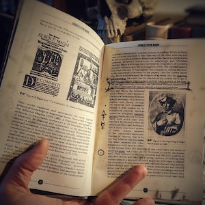 Op de afbeelding: Een open boek met zwarte en witte tekst over alchemie. Het boek is open naar een pagina met een zwart-witte illustratie van een man met een hoed en een pagina met een zwart-witte illustratie van een man in een gewaad. De tekst op de pagina bevat de woorden "De Commixti" en "Albertus Magnus".