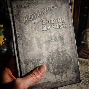 May include: A vintage, worn, black book with the title "Advanced Potion Making" on the cover. The book has a faded image of a cauldron and other potion-making tools.