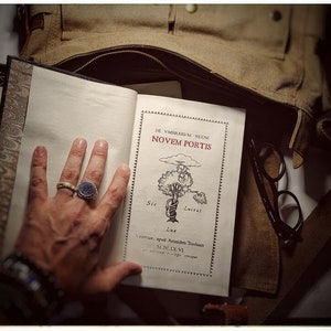 K&ouml;nnte beinhalten: Ein offenes Buch mit einem braunen Lederumschlag und einer Hand, die die Seiten offen h&auml;lt. Das Buch tr&auml;gt den Titel "De Vmbrarvm Regni Novem Portis" und zeigt eine Zeichnung eines Baumes mit einem Seil darum. Das Buch liegt auf einer braunen Stofftasche mit einer Brille darauf.