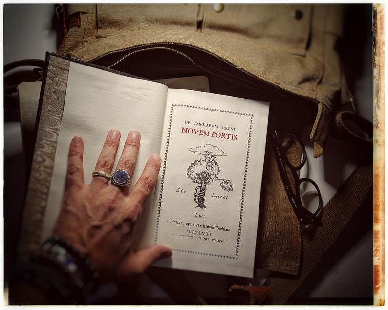 K&ouml;nnte beinhalten: Ein offenes Buch mit einem braunen Lederumschlag und einer Hand, die die Seiten offen h&auml;lt. Das Buch tr&auml;gt den Titel "De Vmbrarvm Regni Novem Portis" und zeigt eine Zeichnung eines Baumes mit einem Seil darum. Das Buch liegt auf einer braunen Stofftasche mit einer Brille darauf.