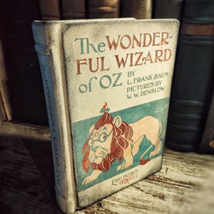 May include: A book titled The Wonderful Wizard of Oz by L. Frank Baum with pictures by W. W. Denslow. The cover features a cream background, teal accents, and a central illustration of a lion with a red mane and glasses. The spine reads the title.