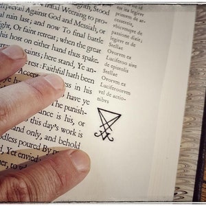 K&ouml;nnte beinhalten: Nahaufnahme eines Buches mit einer Hand, die die Seite offen h&auml;lt. Der Text auf der Seite ist in lateinischer Sprache. Ein schwarzes Pentagramm-Symbol ist unten auf der Seite gezeichnet.