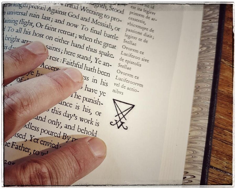 K&ouml;nnte beinhalten: Nahaufnahme eines Buches mit einer Hand, die die Seite offen h&auml;lt. Der Text auf der Seite ist in lateinischer Sprache. Ein schwarzes Pentagramm-Symbol ist unten auf der Seite gezeichnet.