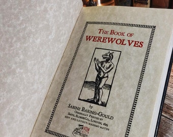 Puede incluir: Un libro antiguo titulado "The Book of Werewolves" de Sabine Baring-Gould. La portada presenta una ilustración en blanco y negro de un hombre lobo de pie sobre sus patas traseras con las manos levantadas en un gesto de oración.