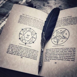 May include: An open book with a black feather quill resting on the pages. The book is titled "The Mysteries of Gothic Theurgy" and "Book of Black Magic". The pages contain handwritten text and diagrams of pentagrams.