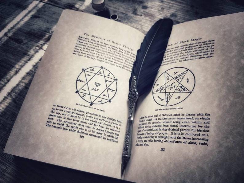 May include: An open book with a black feather quill resting on the pages. The book is titled "The Mysteries of Gothic Theurgy" and "Book of Black Magic". The pages contain handwritten text and diagrams of pentagrams.