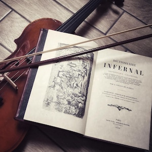 Könnte beinhalten: Ein altes Buch mit dem Titel "Dictionnaire Infernal: Repertoire Universel" von J. Collin de Plancy, aufgeschlagen auf eine Seite mit einer Illustration. Eine Geige und ein Bogen liegen auf dem Buch.