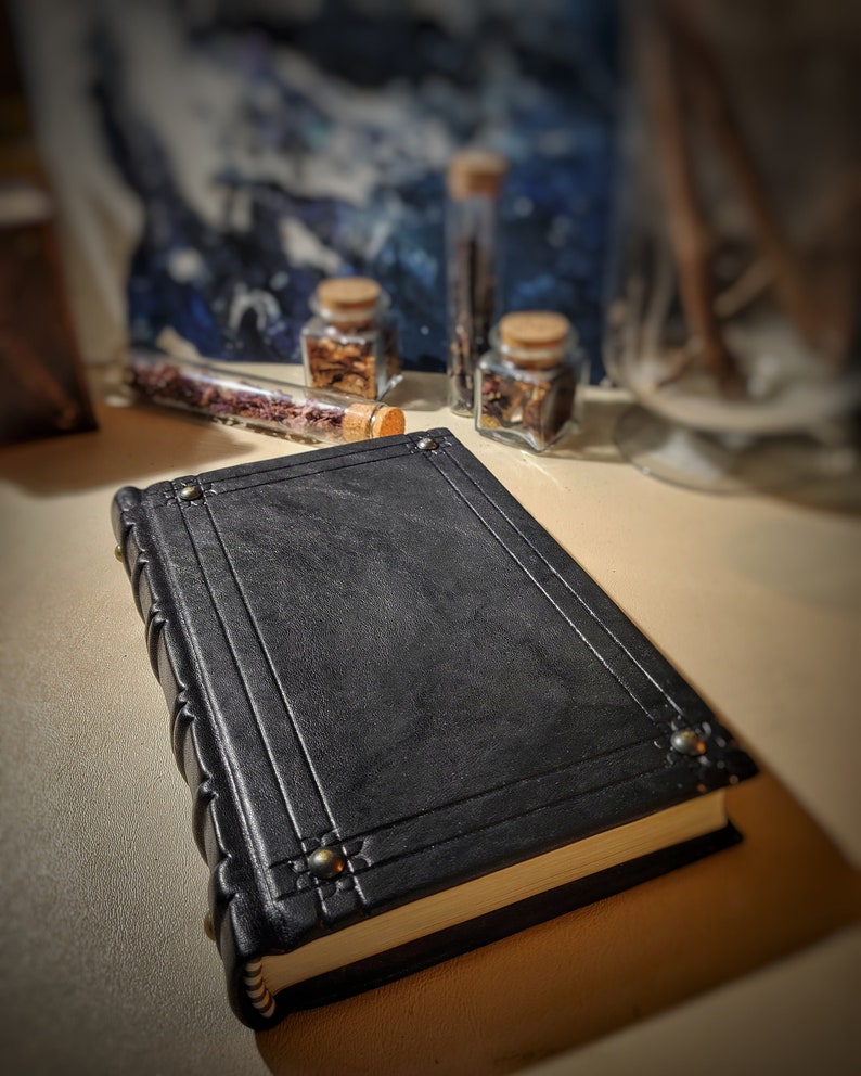 May include: A black leather-bound journal with decorative stitching and brass accents. The journal is closed and lying on a light brown surface.