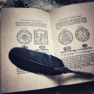 May include: Open book with a black feather quill pen on top of the pages. The book is titled "The Book of Black Magic" and "The Rituals of Black Magic". The pages contain text and illustrations of symbols and sigils.