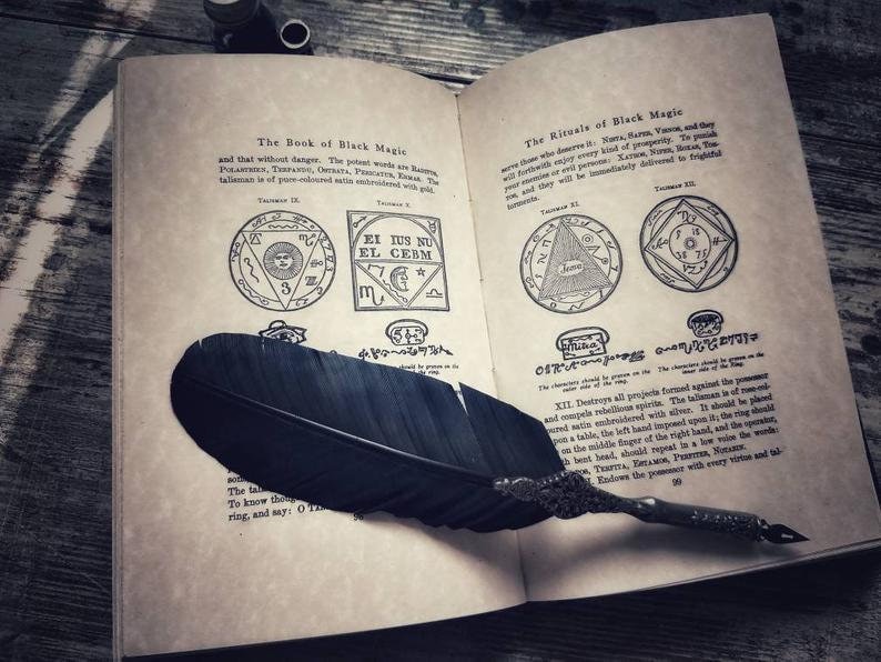 May include: Open book with a black feather quill pen on top of the pages. The book is titled "The Book of Black Magic" and "The Rituals of Black Magic". The pages contain text and illustrations of symbols and sigils.