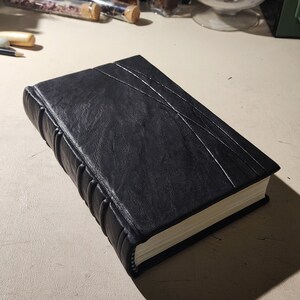 K&ouml;nnte beinhalten: Ein schwarz ledergebundenes Notizbuch mit strukturierter Oberfl&auml;che und dekorativen N&auml;hten. Der Buchr&uuml;cken hat erhabene B&auml;nder, und die Seiten sind wei&szlig; umrandet. Das Notizbuch hat ein klassisches, handgefertigtes Aussehen.