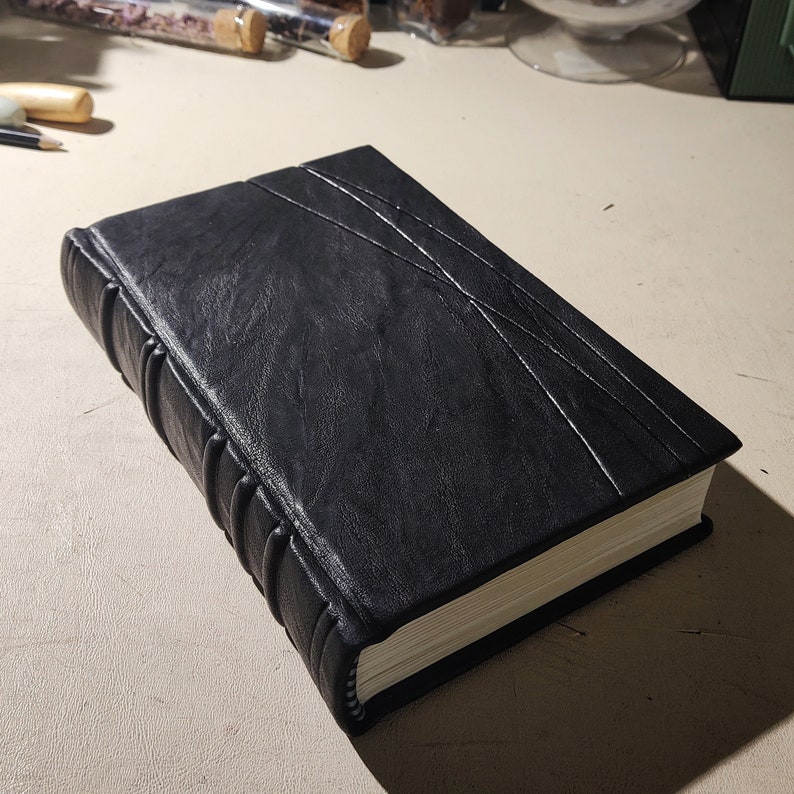 K&ouml;nnte beinhalten: Ein schwarz ledergebundenes Notizbuch mit strukturierter Oberfl&auml;che und dekorativen N&auml;hten. Der Buchr&uuml;cken hat erhabene B&auml;nder, und die Seiten sind wei&szlig; umrandet. Das Notizbuch hat ein klassisches, handgefertigtes Aussehen.