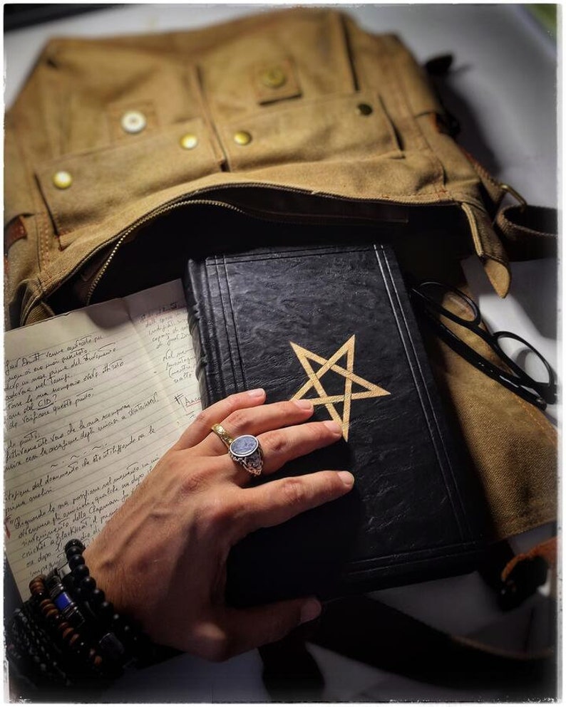 K&ouml;nnte beinhalten: Ein schwarzes, ledergebundenes Tagebuch mit einem goldenen Pentagramm auf dem Einband. Das Tagebuch liegt auf einer braunen Canvas-Tasche und eine Hand h&auml;lt das Tagebuch offen.