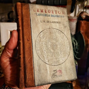 Può includere: Un libro d'epoca intitolato "LEMEGETON CLAVICULA SALOMONIS" di L.W. De Laurence. Il libro ha un aspetto invecchiato e usurato con una copertina chiara con una grafica circolare e rilegatura in pelle marrone. Il libro è tenuto in una mano.