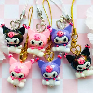 May include: A collection of six character charms featuring Kuromi in various colors including black, pink, and purple. Each charm has a skull emblem on the forehead, pointed ears, and a small heart-shaped metal loop attached to a thin hanging cord.