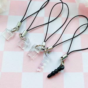 May include: Four phone charm accessories with black cords and silver-colored metal clasps. The charms are clear and black, with a variety of shapes. The background is a pink and white checkered pattern.