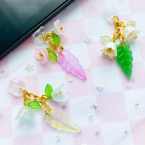 Puede incluir: Tres colgantes decorativos para teléfono con diseños de flores y hojas. Cada colgante tiene una cadena dorada, un accesorio de plástico transparente y acentos coloridos. Las flores son blancas y rosas, y las hojas son verdes y rosas. El fondo es un patrón de cuadros rosas y blancos.