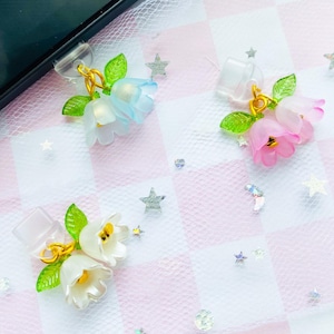 Puede incluir: Se muestran tres colgantes florales para teléfono. Uno es azul claro, otro rosa y otro blanco. Cada colgante tiene un anillo dorado y hojas verdes. El fondo es un patrón de cuadros rosas y blancos con lentejuelas en forma de estrella.