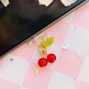 Puede incluir: Un amuleto dorado con dos cerezas rojas y una hoja verde unida a una funda de teléfono.