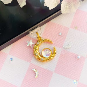 以下が含まれることがあります： 小さな透明なクリスタルと中央に虹色の円形ストーンをあしらった、ゴールドトーンの三日月型チャーム。チャームは透明なプラスチック製の粘着タブに取り付けられており、黒い電子機器の端に固定されています。