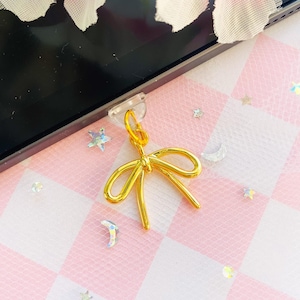 Puede incluir: Un charm de lazo de color dorado con un respaldo adhesivo transparente. El charm está sobre un fondo a cuadros rosa y blanco con purpurina.