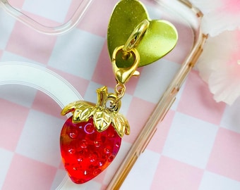 Può includere: Un ciondolo a forma di fragola rossa traslucida con una parte superiore in metallo dorato a forma di foglia, fissato a un moschettone dorato e a un piccolo supporto in metallo verde a forma di cuore, come accessorio decorativo per una cover trasparente.