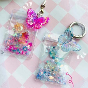 May include: Two clear, rectangular pouches with butterfly charms and customizable fillings. One pouch has a bright pink butterfly, the other a light blue butterfly. The pouches are filled with colorful stars, beads, and glitter. Text reads "CUSTOMIZABLE FILLING".