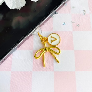 May include: A gold-colored phone charm featuring a bow design and a circular charm with the letter "A" in white. The charm is attached to a phone with a clear adhesive. The background is a pink and white checkered pattern.