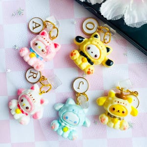 May include: A collection of colorful cartoon character charms. The charms feature various characters in pink, yellow, white, and blue, each with a unique design. They are attached to gold-colored keychains with clear plastic clips.