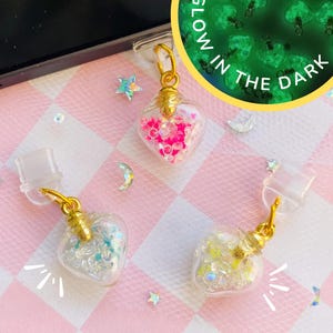 以下が含まれることがあります： ゴールドトーンの金属製キャップと透明なプラスチックループが付いた、ハート型のガラス瓶チャーム3個。瓶の中には、小さな虹色とピンクの装飾ビーズが入っています。円形のインセット画像には、緑色に光るチャームと「GLOW IN THE DARK」という文字が表示されています。