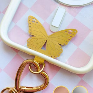 Könnte beinhalten: Ein goldfarbener Schmetterlingsanhänger mit filigranen Ausschnitten ist an einer weißen Handyhülle befestigt. Ein goldfarbener herzförmiger Verschluss und ein weißer rechteckiger Handygriff sind ebenfalls sichtbar. Der Hintergrund zeigt ein rosa-weißes Karomuster.