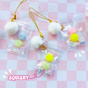 Könnte beinhalten: Transparente Plastikbeutel mit bunten Inhalten, darunter Glitzer, Pompons und kleine, pastellfarbene Formen. Jeder Beutel hat ein goldfarbenes Schlüsselband und einen flauschigen weißen Pompon. Das Wort "SQUISHY" ist rosa geschrieben.
