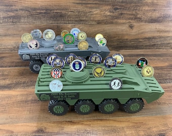 May include: Two toy military vehicles, one gray and one green, are covered with collectible coins. The coins feature various military and patriotic designs. The vehicles are on a wooden surface.