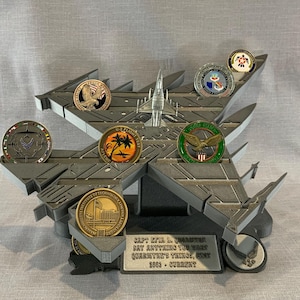 May include: A gray model of an F-16 fighter jet with a display stand. The stand has a plaque that reads "Capt. Eric L. Quarante, Say Anything for What Country's Things, 2003 - Current". The model has several small, round, metal coins in the slots on the wings and fuselage.