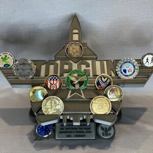 May include: A wooden display stand shaped like a fighter jet with the words "TOP GUN" on the front. The stand has a variety of military challenge coins displayed on it. The stand also has a plaque that reads "GOT ROOM FOR ANYTHING YOU WANT.  QUADRANT THREE, KTY. 2021 - CURRENT."