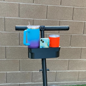 May include: A black scooter handlebar with a built-in cup holder. The holder has two blue and purple tumblers and a phone in a patterned case.