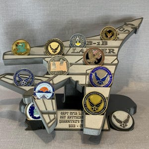 May include: A gray 3D printed B-1B Lancer model with a variety of military challenge coins displayed on it. The coins feature various military insignia and designs.