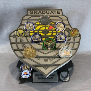 May include: A gray wooden coin display with the words "Graduate", "US Air Force", and "Weapons School" on it. The display has 12 slots for coins, and each slot is filled with a different coin. The coins are all silver or gold in color, and they have various designs on them.
