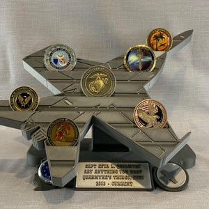 May include: A gray, jet-shaped display stand holding several military challenge coins. The coins feature various emblems and designs, including an eagle, a palm tree, and a Marine Corps emblem. The stand has a plaque with text.
