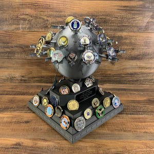 May include: A dark gray globe-shaped display stand holding numerous military challenge coins. The stand has a tiered base with additional coins and a plaque. The coins feature various emblems and colors, creating a detailed and commemorative piece.