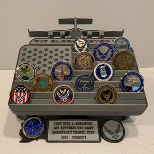 May include: A gray 3D printed display stand with a C-17 Globemaster III airplane on top. The stand has slots for holding 18 challenge coins, and a small clock and a plaque with the text "CAPT EFIA L. QUARMYNE SAY ANYTHING YOU WANT QUARMYNE'S THINGS, ETSY 2003 - CURRENT" are attached to the bottom.