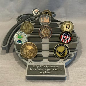 May include: A gray, 3D printed, grenade-shaped coin display stand with a personalized inscription: "MSgt Efia Quarmyne Say whatever you want to say here!" The stand holds a variety of military and other themed coins.