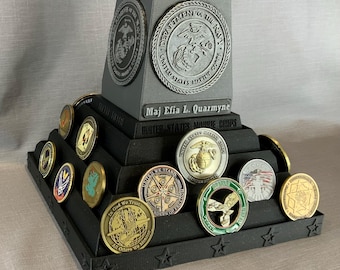 Op de afbeelding: Een zwarte, gelaagde displaystandaard met een grijs, gegraveerd embleem van het United States Marine Corps bovenop. De standaard is gevuld met verschillende militaire challenge-munten, waaronder een gouden munt met de woorden "United We Stand" en een groene munt met de woorden "In God We Trust".
