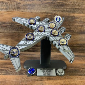 May include: A gray airplane-shaped display stand holding several military challenge coins. The coins have blue and gold detailing. The stand has a black base and a plaque with text. The background is a wooden surface.